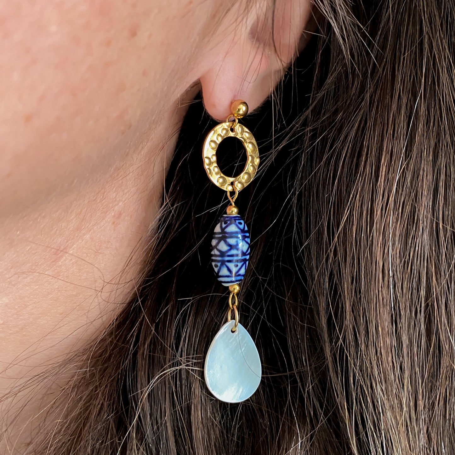 Boucles d’oreilles Abalone portées en gros plan, avec cercle doré martelé, perle ovale en céramique bleue et pendentif en nacre naturelle en forme de goutte.