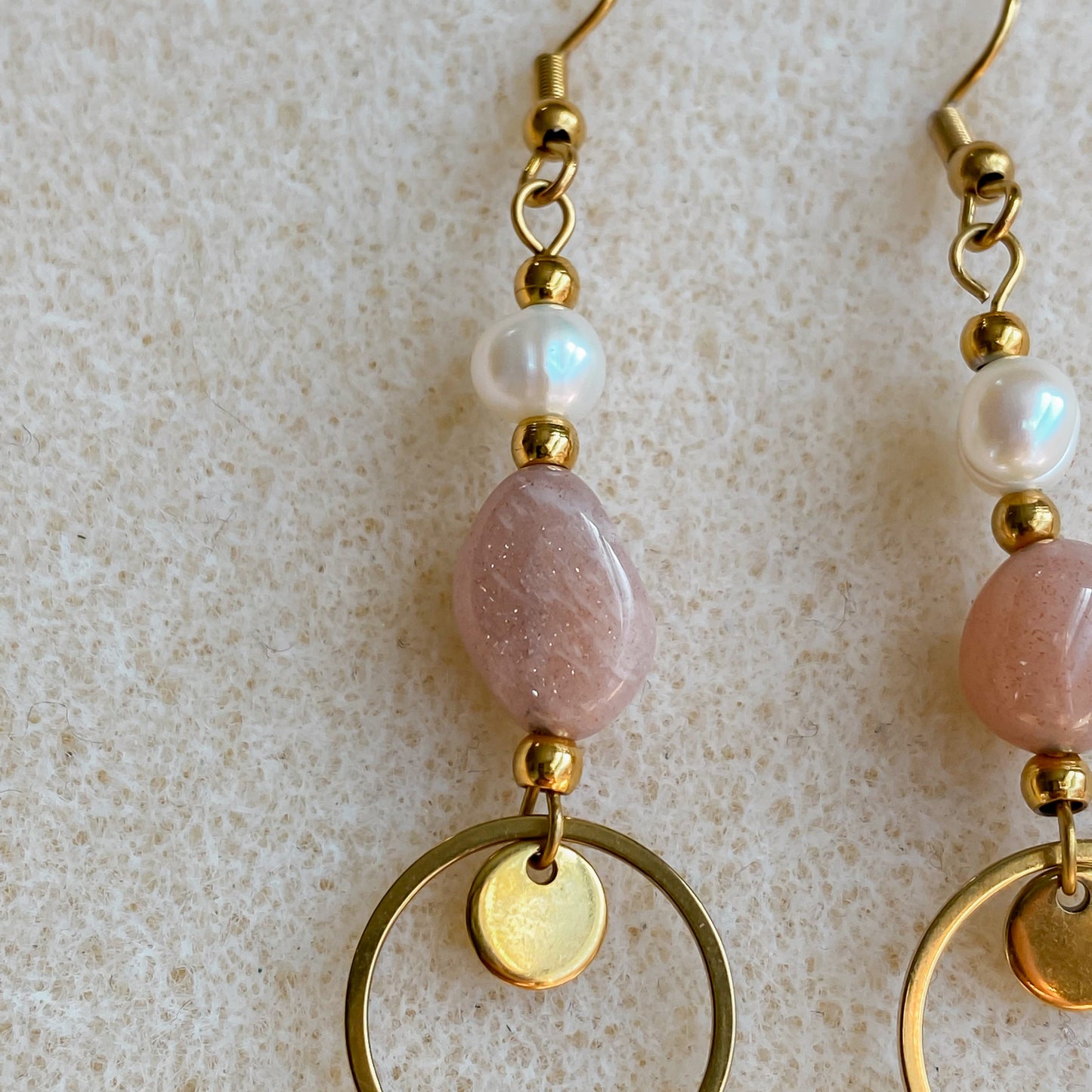 Gros plan sur une boucle Daphné montrant la pierre de soleil rose, la perle d’eau douce et la breloque cercle dorée sur fond texturé beige