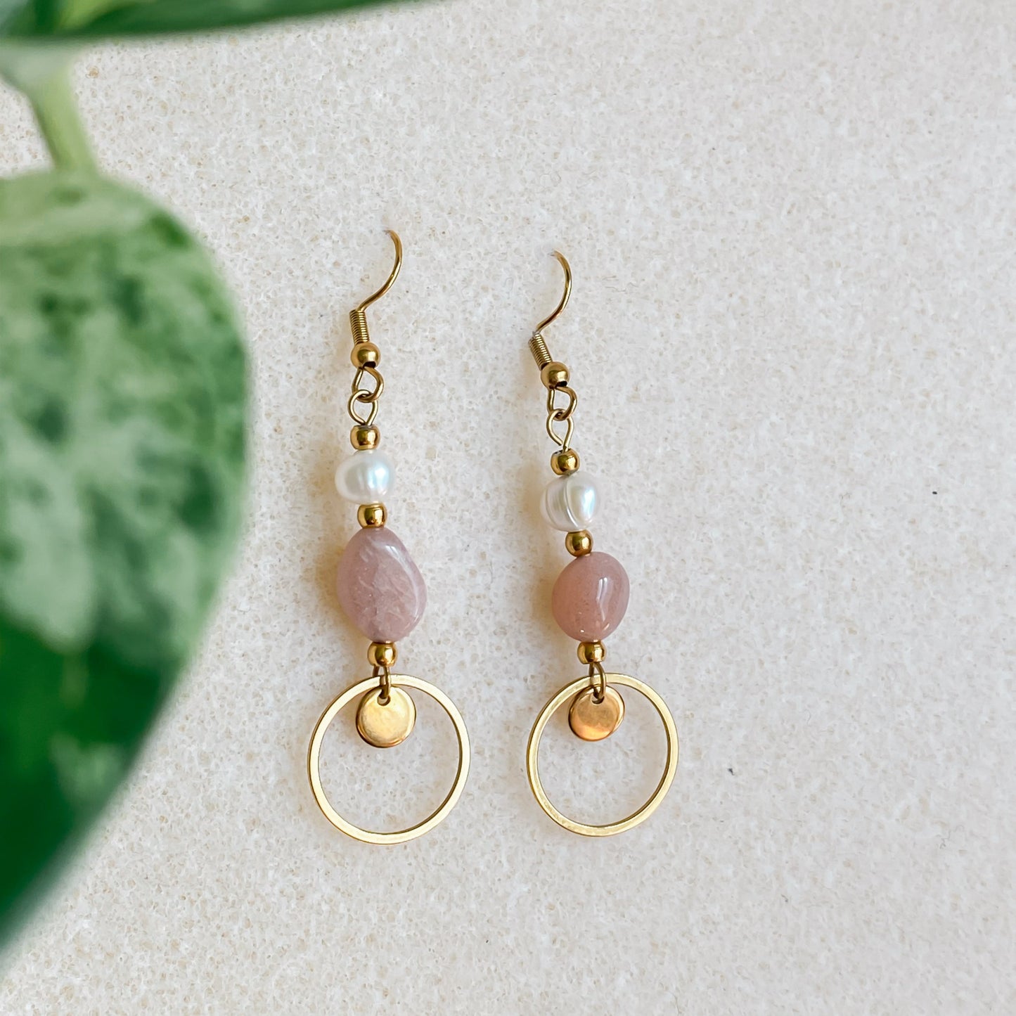 Boucles d’oreilles pendantes Daphné en pierre de soleil et perles d’eau douce, présentées en flatlay sur fond beige avec feuille de plante verte