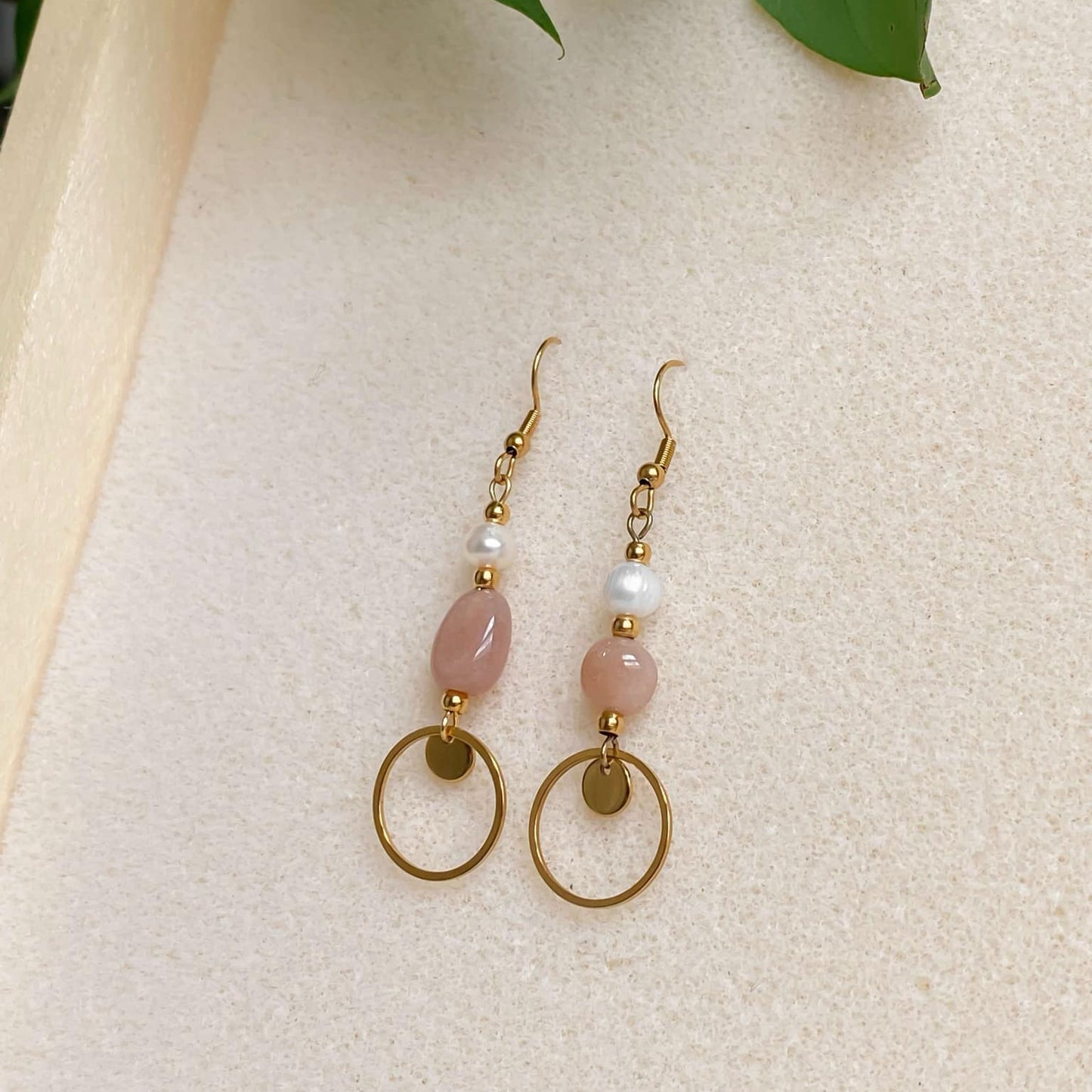 Boucles d’oreilles pendantes Daphné en plaqué or avec perles d’eau douce et pierre de soleil, présentées en flatlay sur fond en mousse beige avec feuilles de plante décoratives