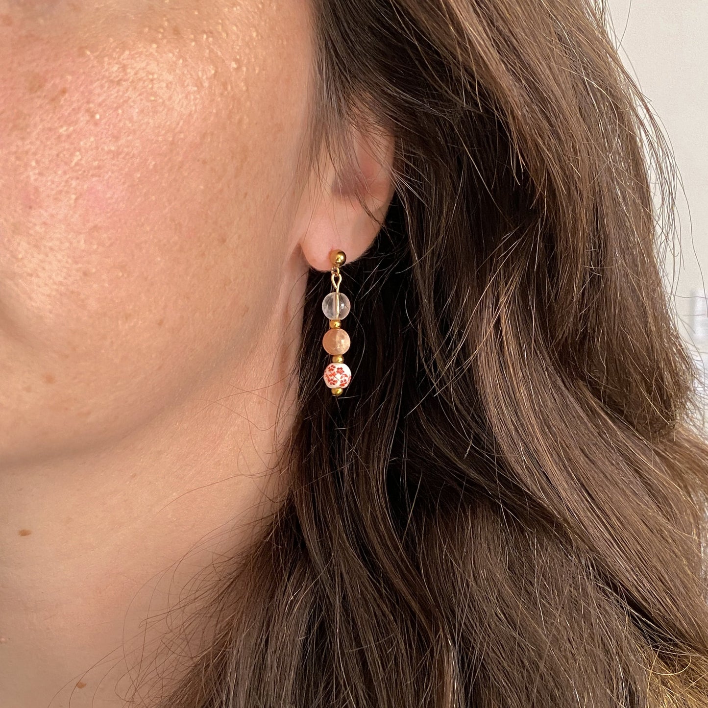 Boucles d’oreilles en pierres naturelles Hanami portées à l’oreille, avec quartz clair, pierre de soleil et bille de céramique florale rouge