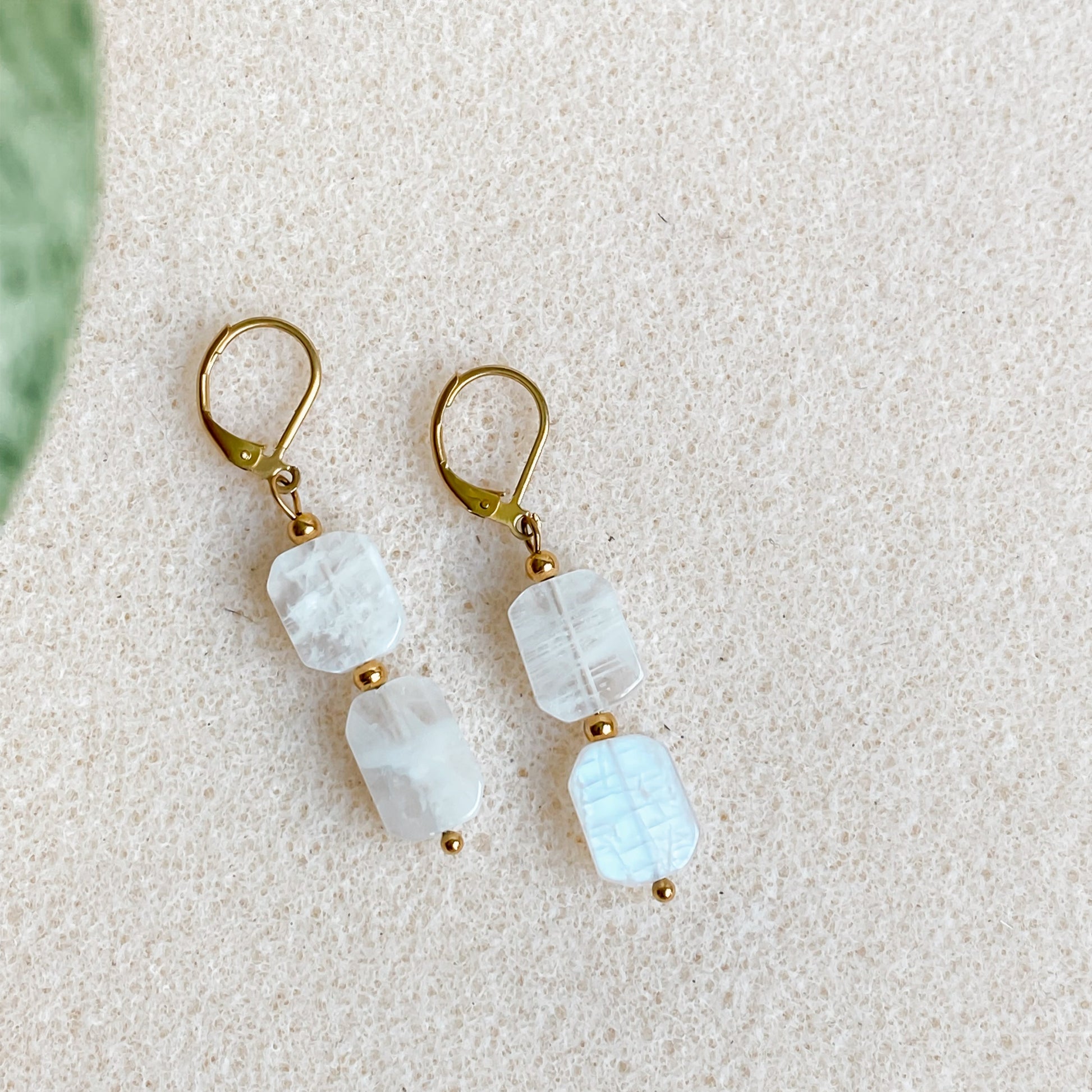 Boucles d’oreilles pendantes Phoebe en pierre de lune et acier inoxydable doré 18K, présentées sur fond beige clair.