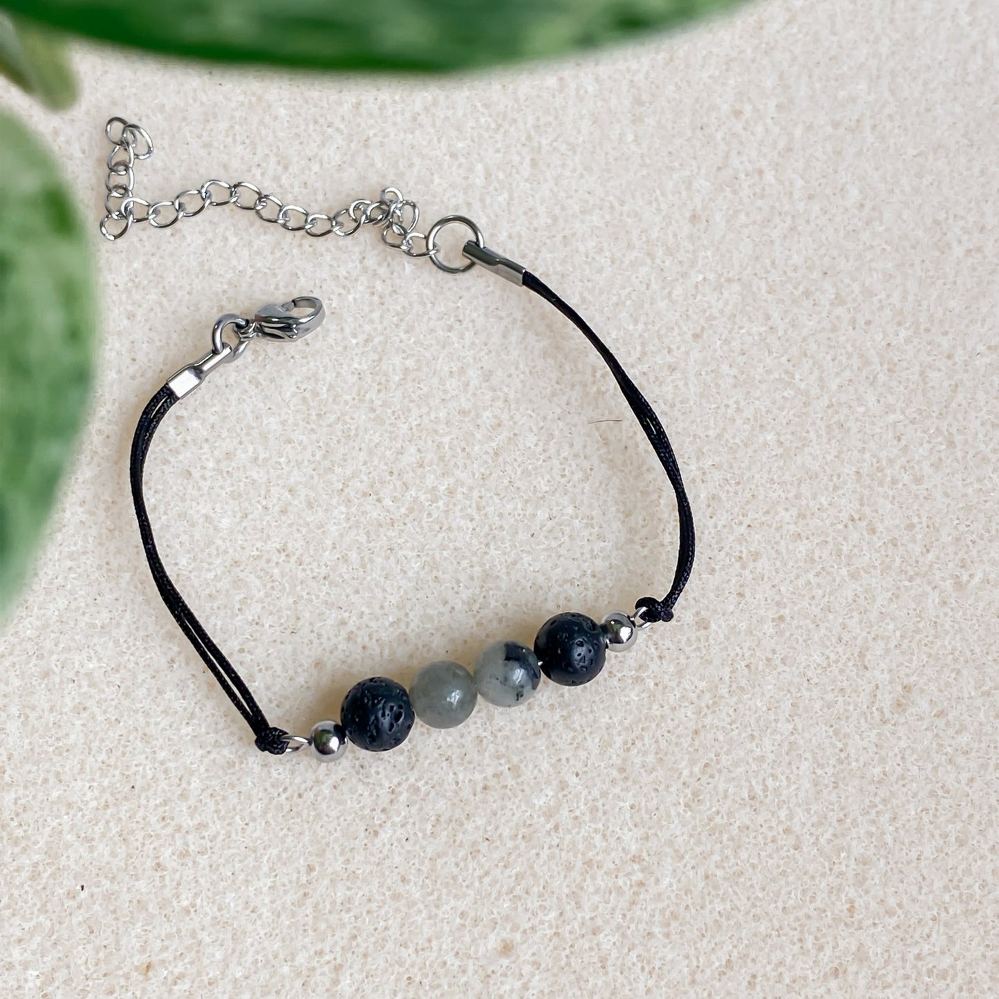 Bracelet ajustable en pierre volcanique et labradorite, cordon noir et fermoir argent, posé sur une surface texturée beige.