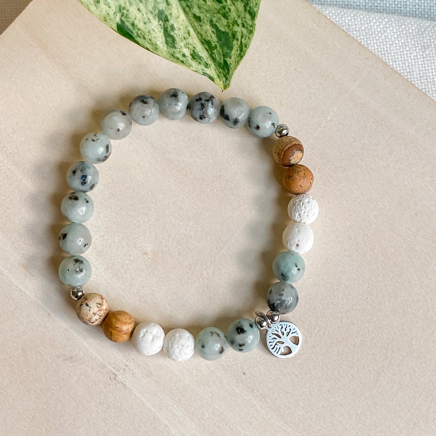 Bracelet arbre de vie en pierres naturelles jaspe kiwi, jaspe paysage et lave blanche, avec pendentif argenté, posé sur bois clair.