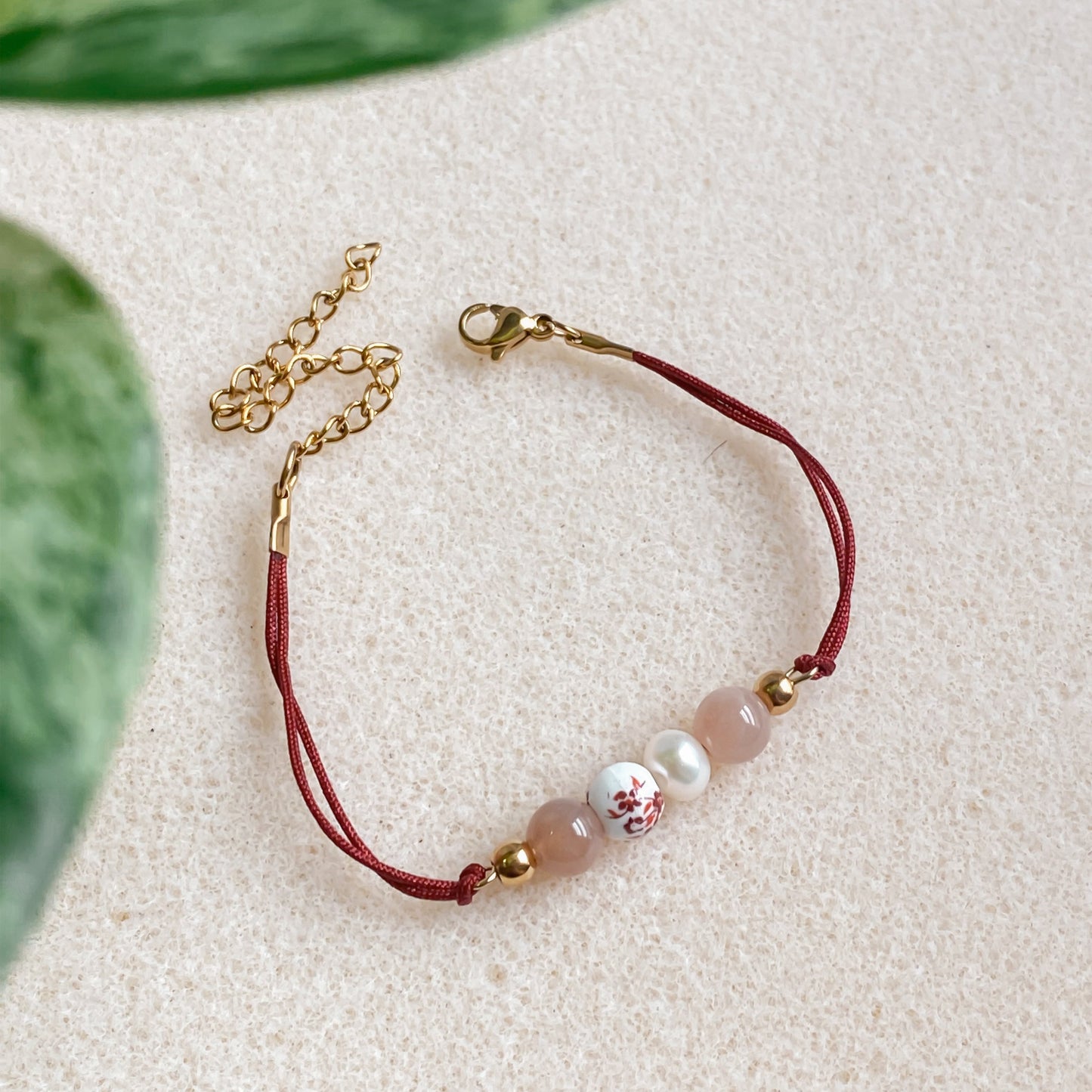 Bracelet cordon rouge vin ajustable Éveil avec pierre de soleil, perle d’eau douce et céramique fleurie, fermoir mousqueton doré et chaîne d’extension.