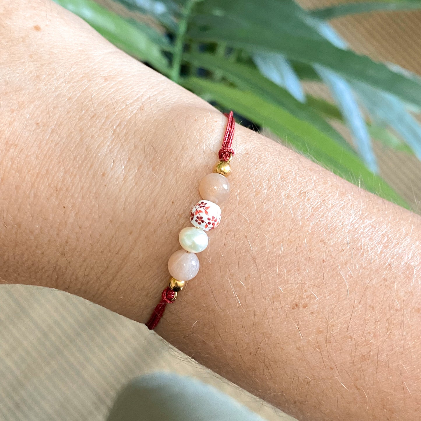 Bracelet cordon rouge en pierres naturelles Éveil porté au poignet, bijou artisanal avec pierre de soleil, perle d’eau douce et céramique fleurie.