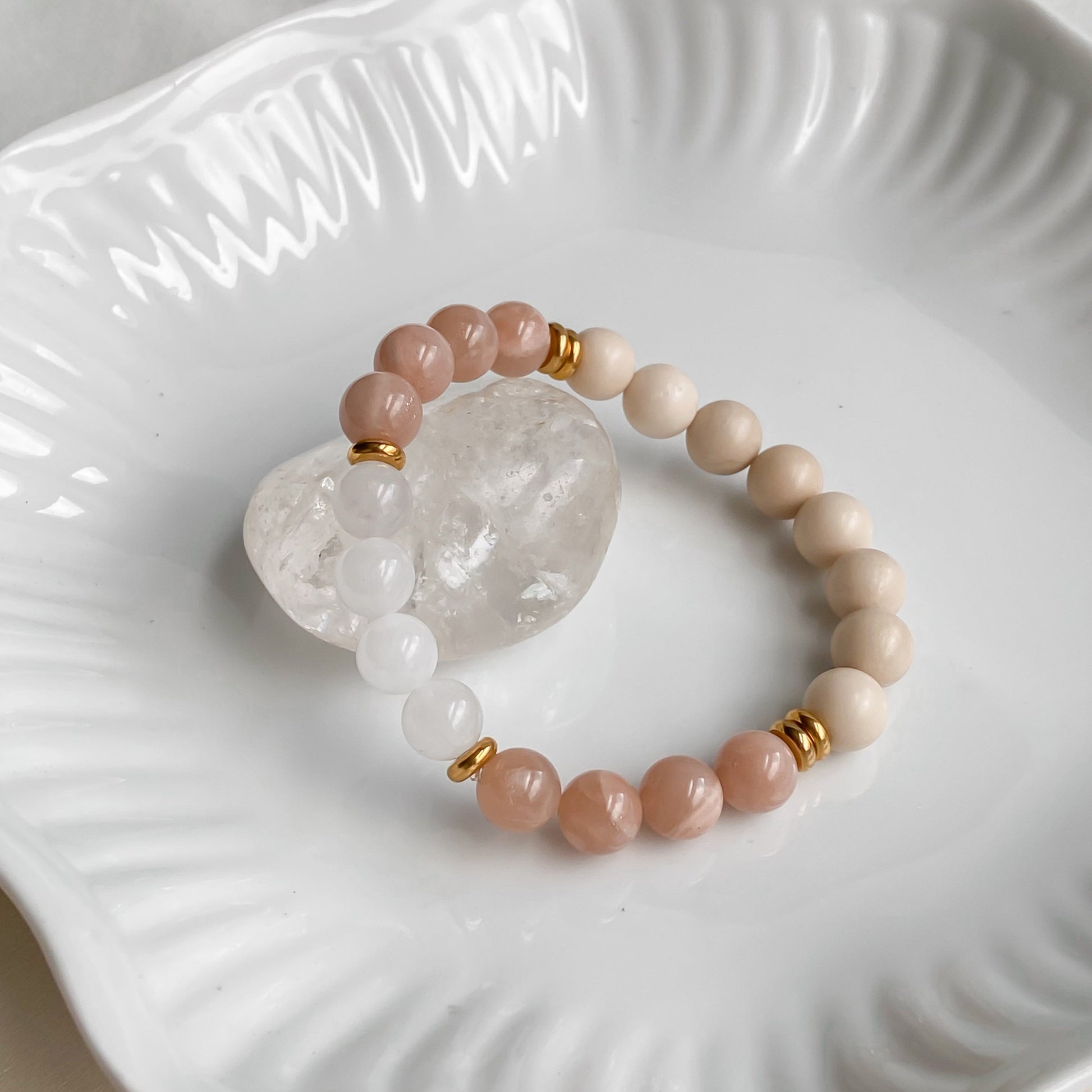 Bracelet Éclat de Soleil en pierre de soleil, pierre de rivière et quartz blanc 8mm avec acier inoxydable doré, vu sous un angle légèrement en hauteur, posé sur une pierre brute de quartz clair dans une assiette blanche sur une planche de bois clair et une nappe en coton naturel.