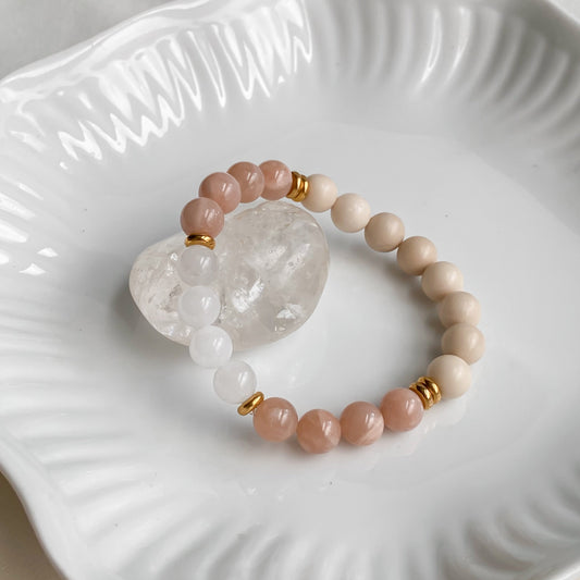Bracelet Éclat de Soleil en pierre de soleil, pierre de rivière et quartz blanc 8mm avec acier inoxydable doré, vu sous un angle légèrement en hauteur, posé sur une pierre brute de quartz clair dans une assiette blanche sur une planche de bois clair et une nappe en coton naturel.