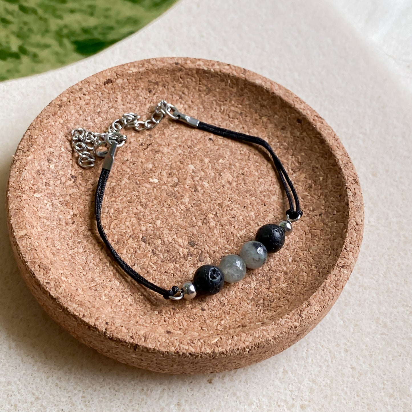 Bracelet minimaliste homme en pierre de lave et labradorite sur cordon noir ajustable, vu de dessus posé sur un support en liège.