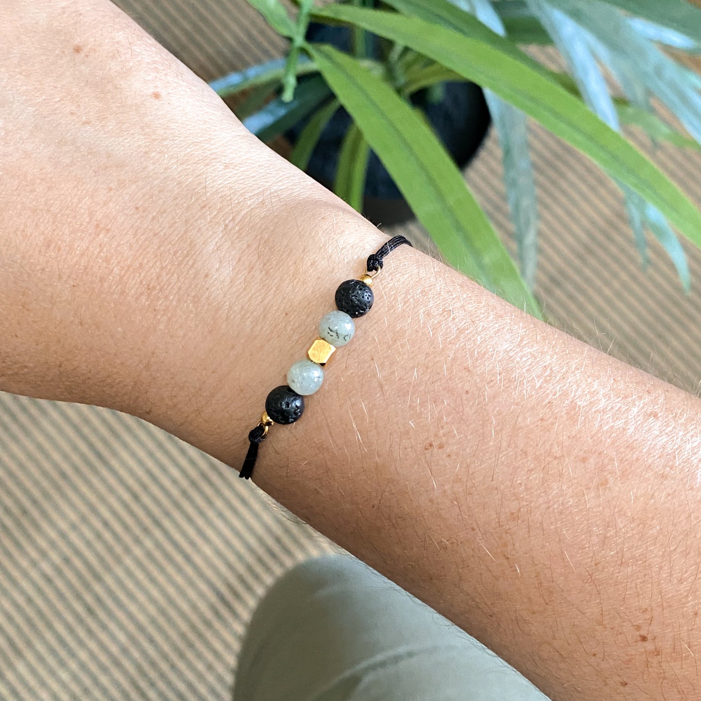 Bracelet NOCTIS pour aromathérapie en pierre de lave et labradorite, cordon noir fin et ajustable, porté au poignet avec plante verte en arrière-plan.