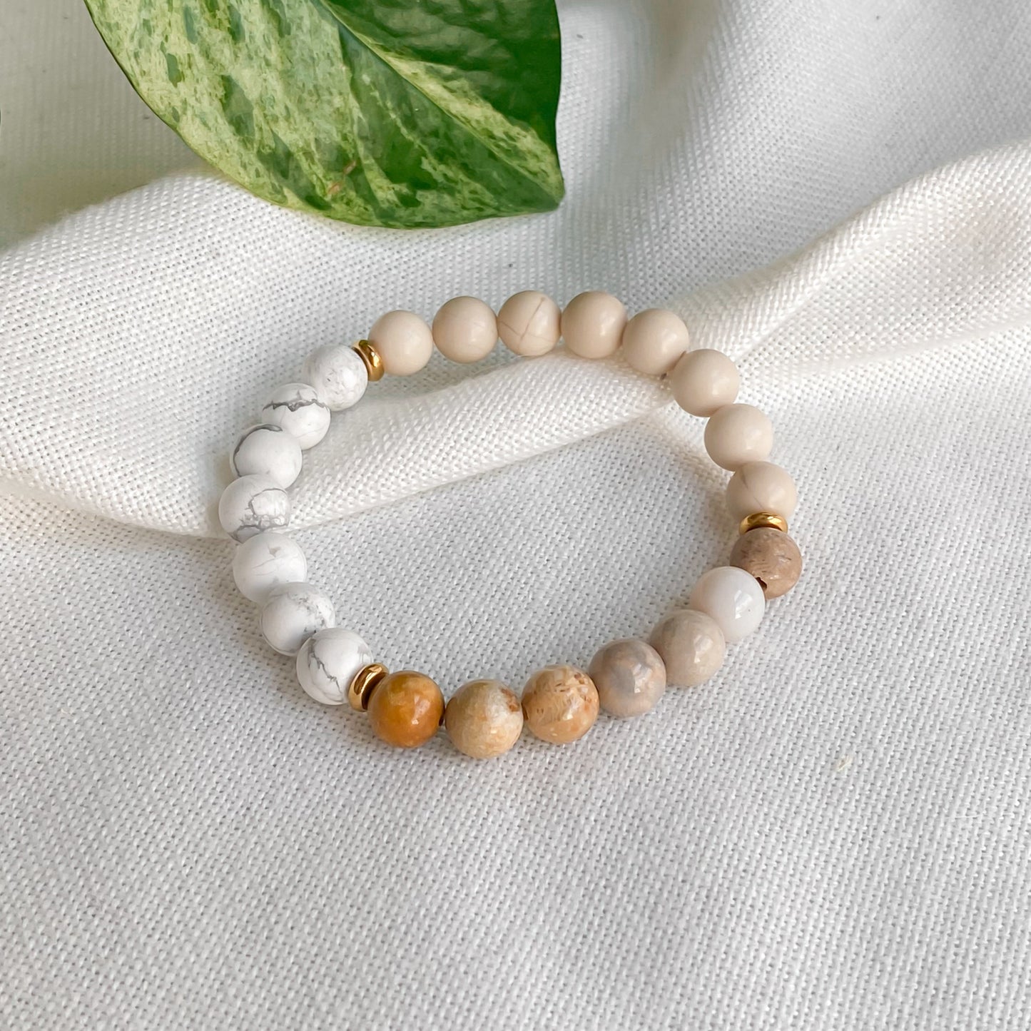 Bracelet élastique Renouveau en pierres naturelles 8mm (jaspe chrysanthème, pierre de rivière, howlite) et acier inoxydable doré, vu sous un léger angle pour mieux apprécier les détails des pierres, sur une nappe en coton naturel avec des plantes.