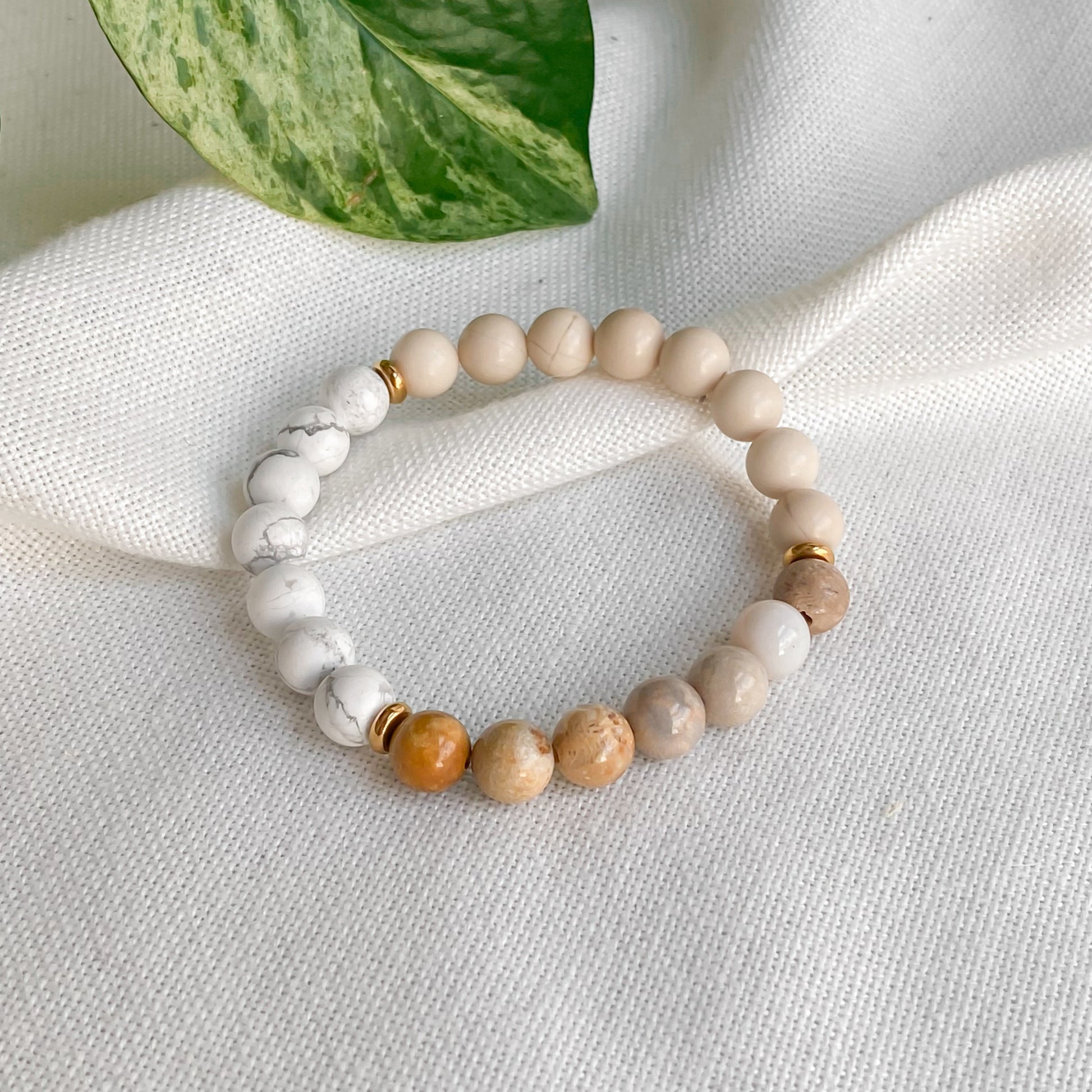 Bracelet élastique Renouveau en pierres naturelles 8mm (jaspe chrysanthème, pierre de rivière, howlite) et acier inoxydable doré, vu sous un léger angle pour mieux apprécier les détails des pierres, sur une nappe en coton naturel avec des plantes.