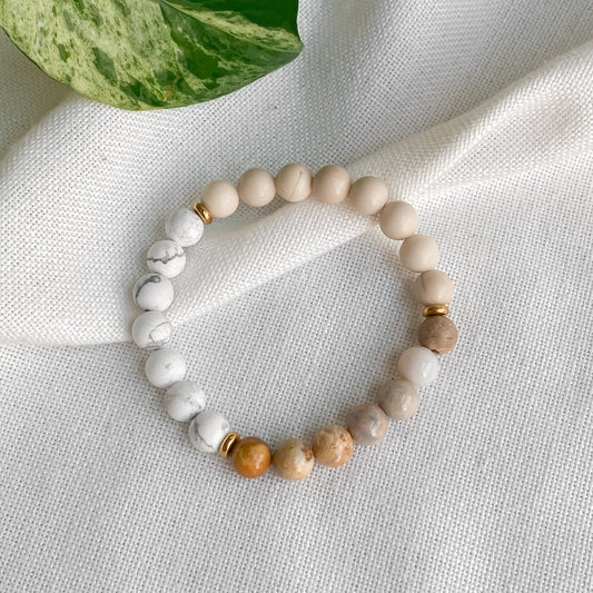 Bracelet élastique Renouveau en jaspe chrysanthème, pierre de rivière et howlite (8mm) avec acier inoxydable doré, présenté sur une nappe en coton naturel entouré de plantes.