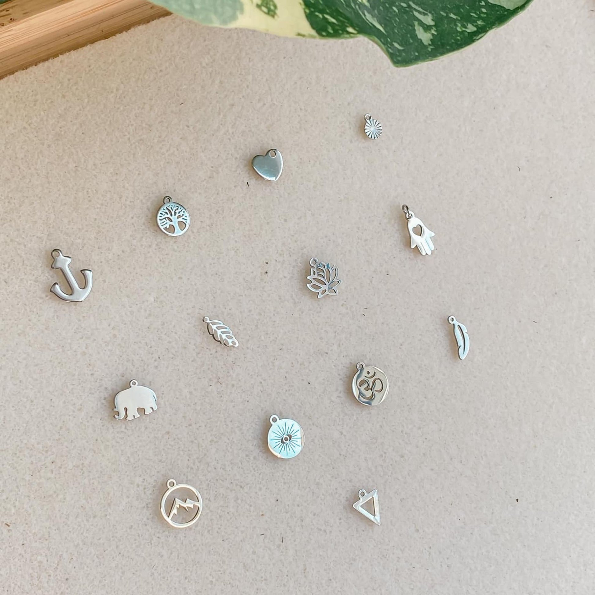 Gros plan sur les breloques en acier inoxydable argenté pour bracelets cordon personnalisables, mettant en valeur les motifs détaillés comme ancre, arbre de vie, cœur, disque, éléphant, feuille, fleur de lotus, main de Fatma, montagnes, œil protecteur, ohm, plume, et triangle.