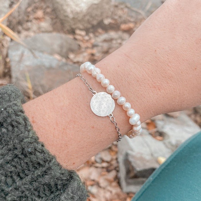 Bracelet minimaliste en chaîne fine avec médaillon fleur de vie, porté au poignet avec un bracelet en perles d'eau douce pour un look élégant et naturel.