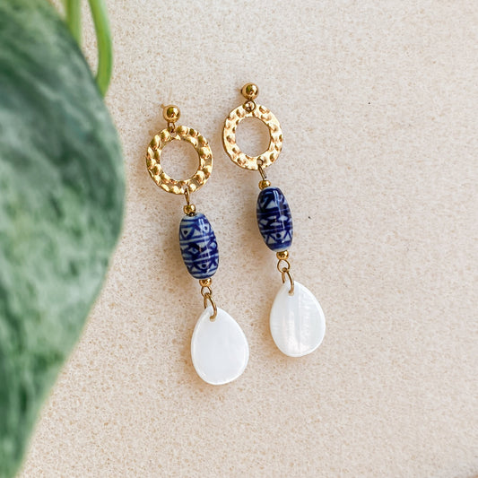 Boucles d’oreilles Abalone en flatlay sur fond beige avec feuille verte, mélange de doré, céramique bleue et nacre naturelle.