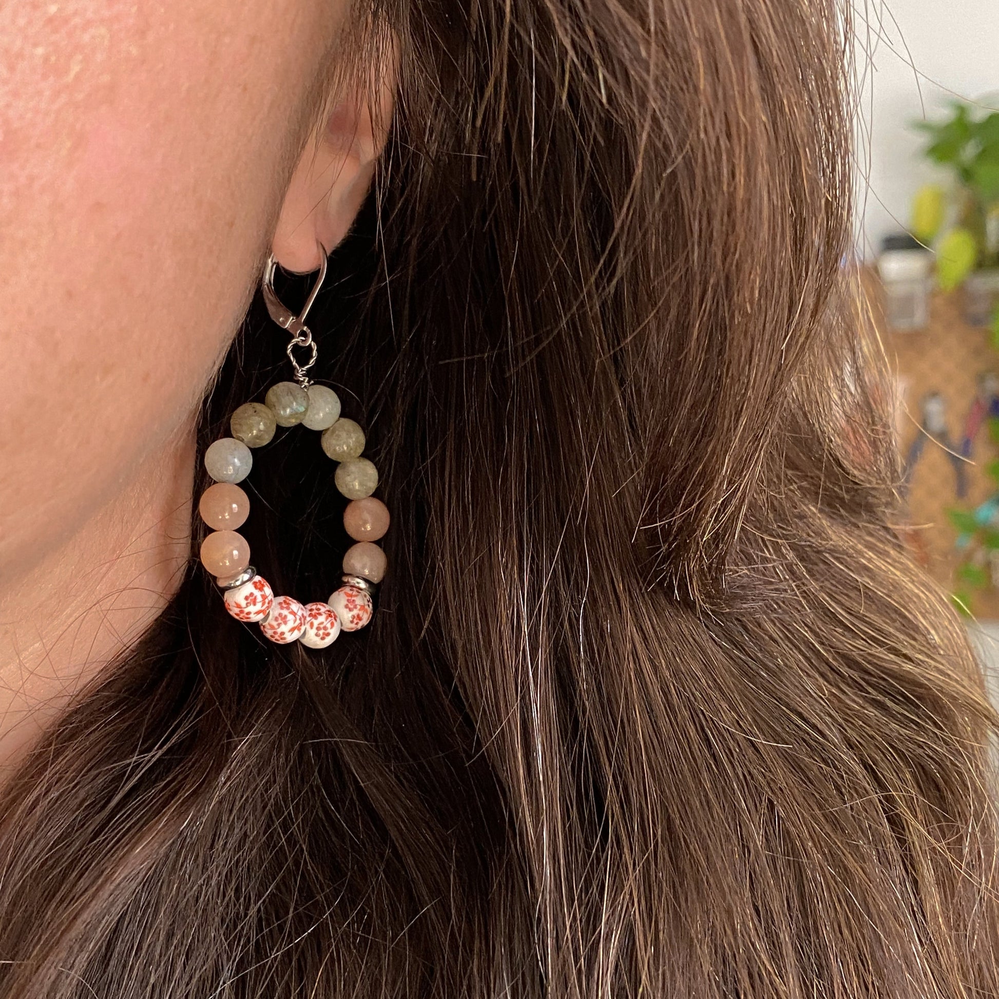 Boucle d’oreille pendante Capri portée à l’oreille, en labradorite, pierre de soleil et céramique fleurie rouge – bijou fait main à Montréal.