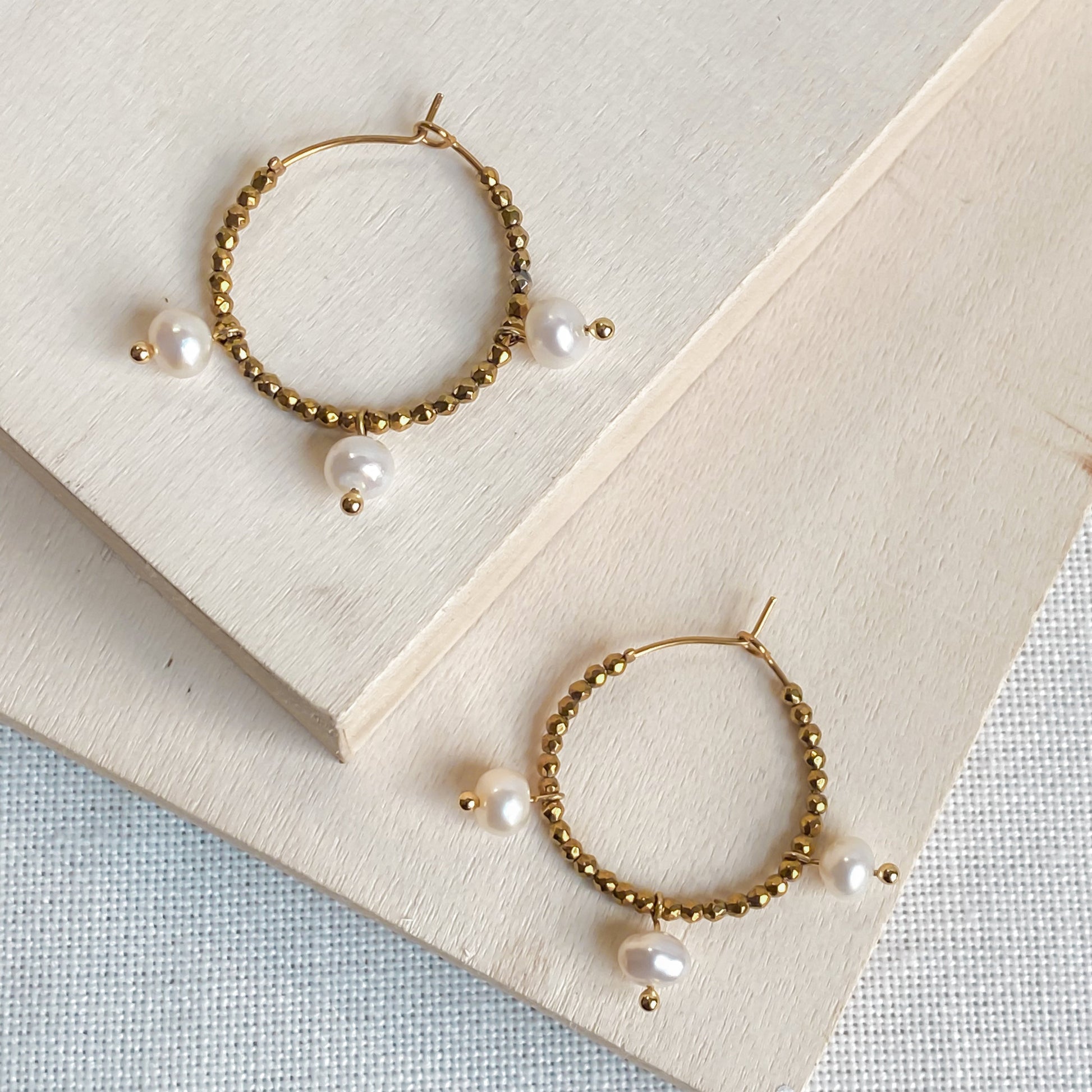 Paire de boucles d’oreilles Auréa, créoles dorées artisanales avec gouttes nacrées d’eau douce, posées sur planche de bois.