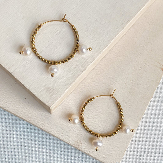 Paire de boucles d’oreilles Auréa, créoles dorées artisanales avec gouttes nacrées d’eau douce, posées sur planche de bois.