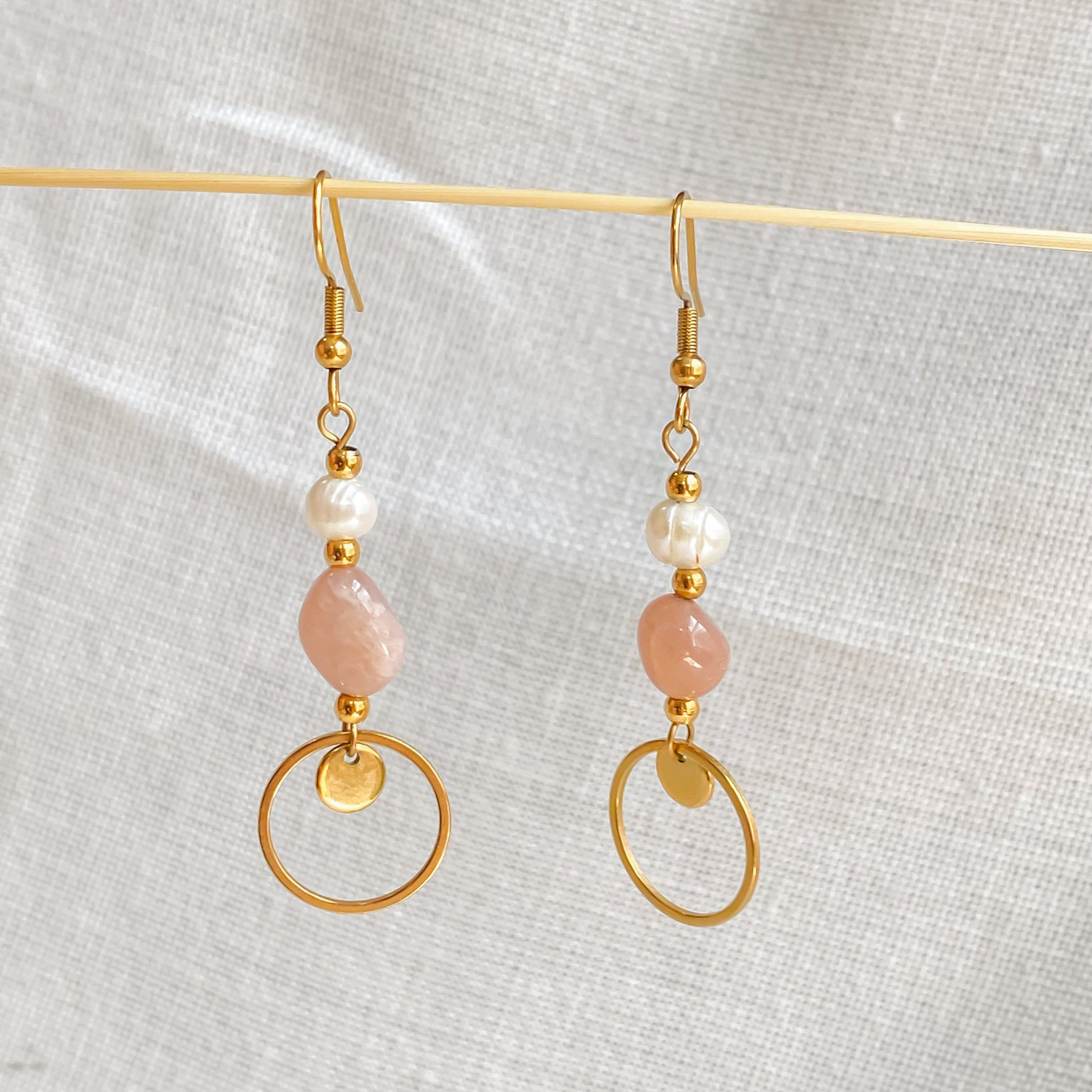 Boucles pendantes Daphné en acier inoxydable plaqué or, avec perles d’eau douce et pierre de soleil, photographiées sur fond tissu beige clair