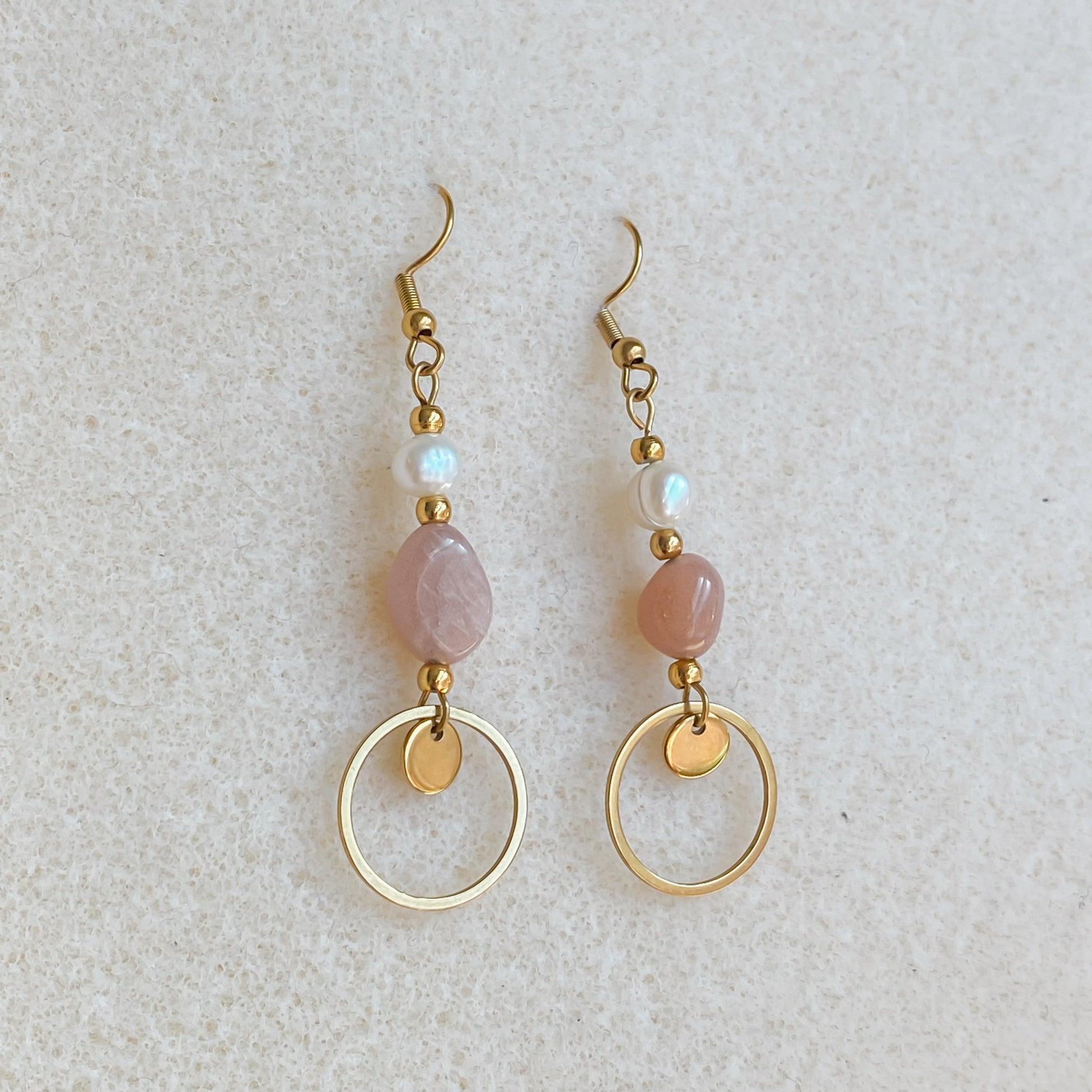 Boucles d’oreilles pendantes Daphné en plaqué or avec perles d’eau douce et pierre de soleil, photographiées en flatlay sur fond beige texturé