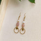 Boucles d’oreilles pendantes Daphné en plaqué or avec perles d’eau douce et pierre de soleil, présentées en flatlay sur fond en mousse beige avec feuilles de plante décoratives