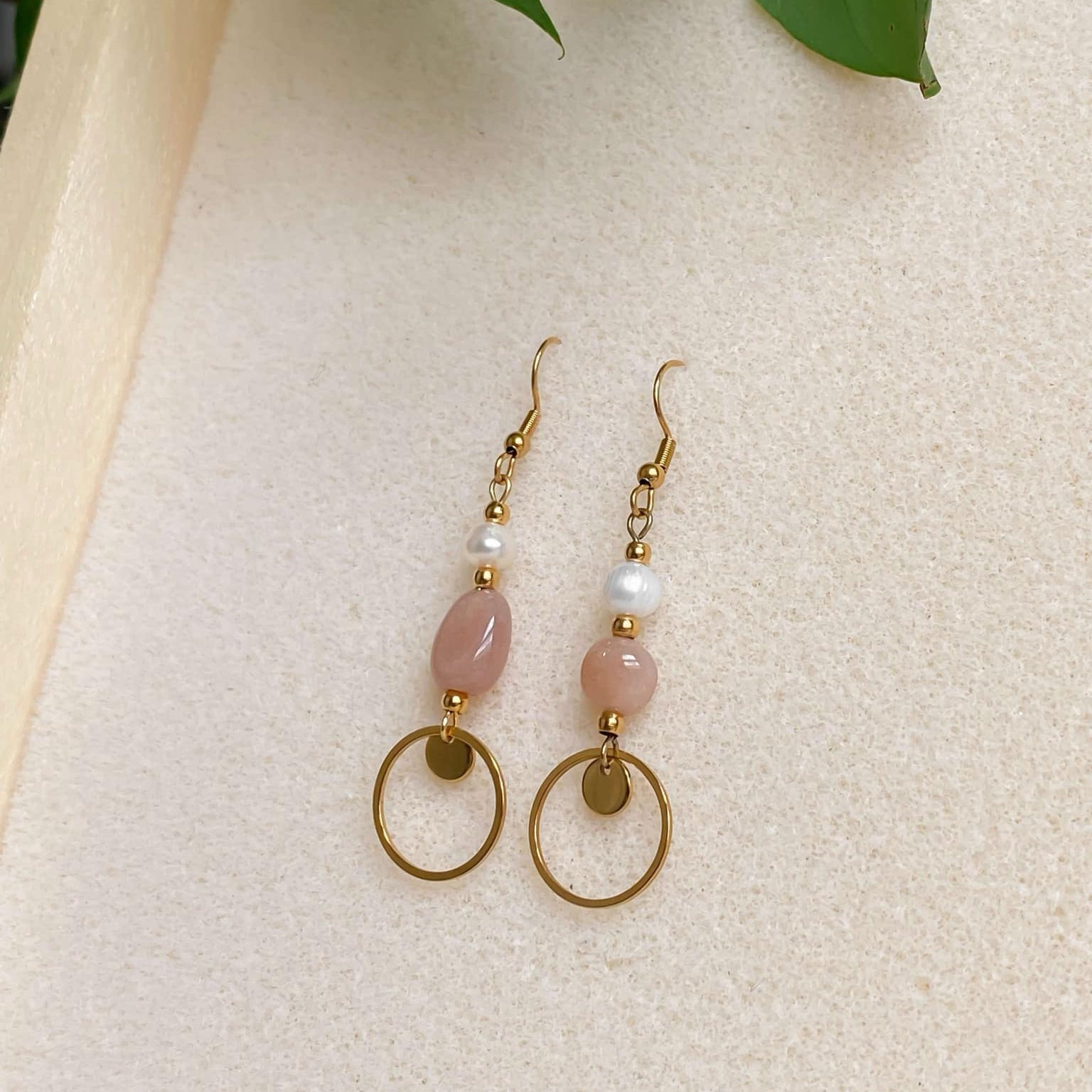 Boucles d’oreilles pendantes Daphné en plaqué or avec perles d’eau douce et pierre de soleil, présentées en flatlay sur fond en mousse beige avec feuilles de plante décoratives
