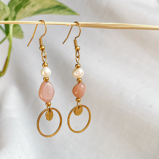 Boucles d’oreilles pendantes Daphné en plaqué or avec perles d’eau douce et pierre de soleil, suspendues sur fond blanc avec feuille verte floutée