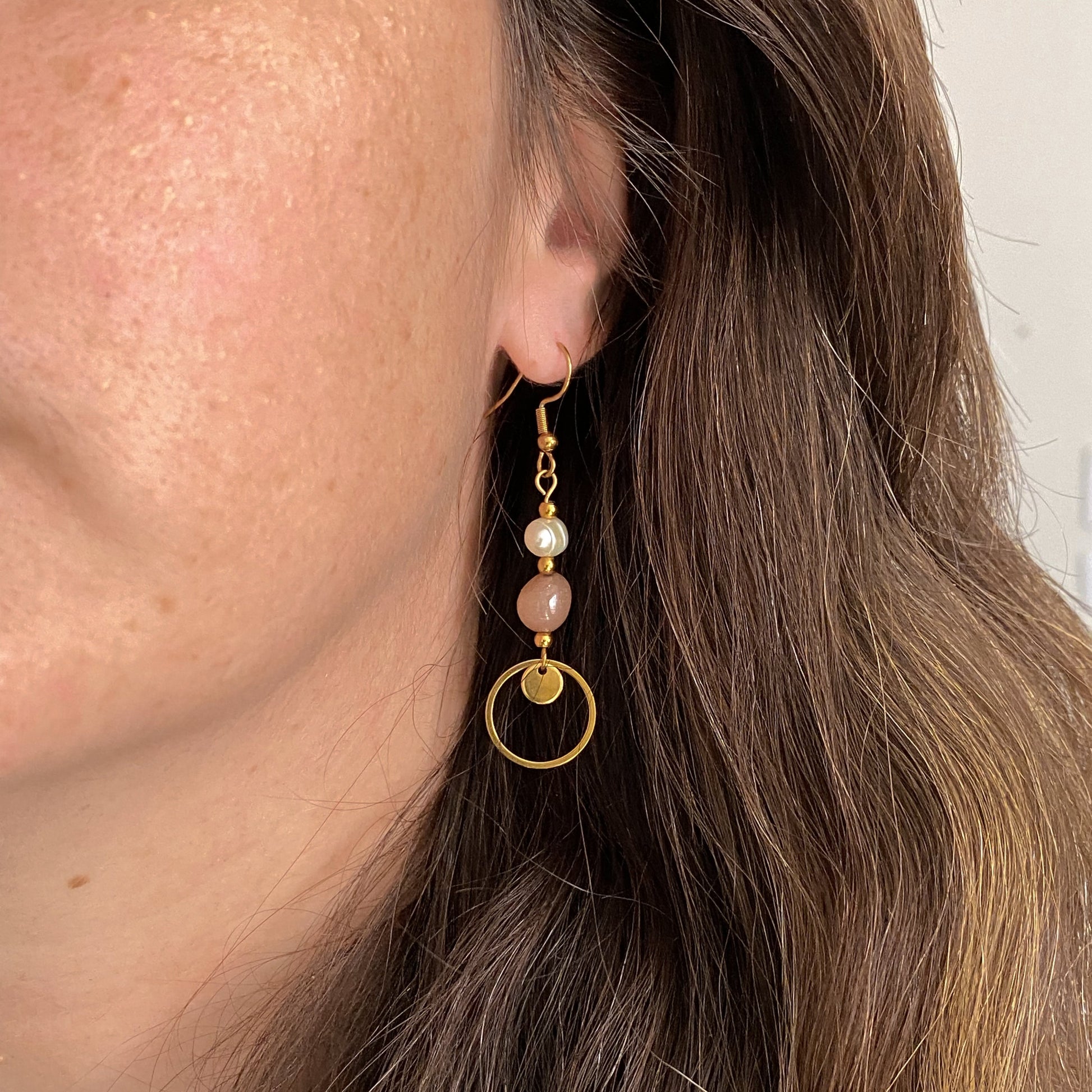 Boucles pendantes Daphné en pierre de soleil et perle d’eau douce avec breloque cercle dorée, portées à l’oreille pour montrer la longueur et le style