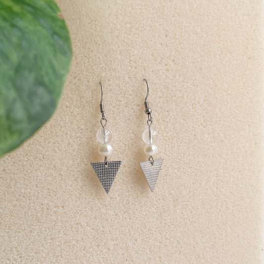 Boucles d'oreilles pendantes Perlie en acier inoxydable avec perles d'eau douce et quartz clair, vue de face, sur fond texturé blanc cassé et feuille de plantes.