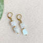 Boucles d’oreilles pendantes Phoebe en pierre de lune et acier inoxydable doré 18K, présentées sur fond beige clair.