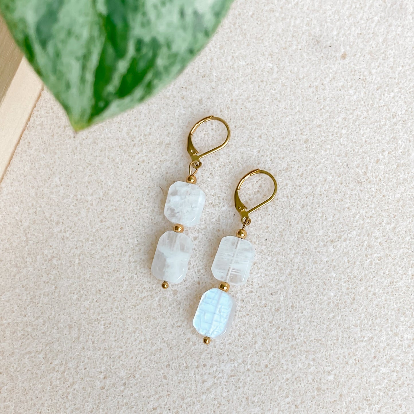 Boucles Phoebe avec pierres de lune rectangulaires irrégulières et fermoir doré, posées sur fond minéral avec feuille verte.