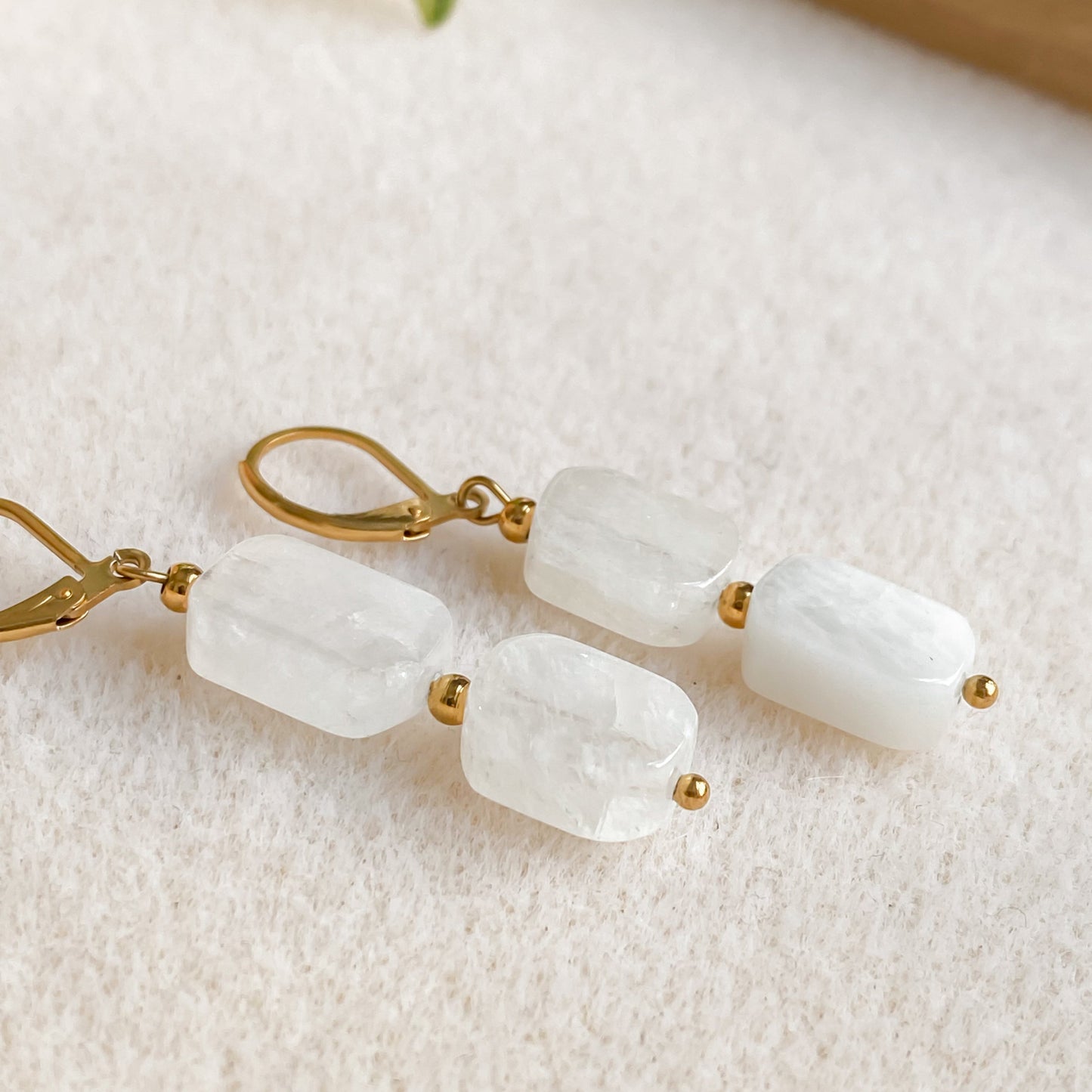 Boucles Phoebe en pierre de lune aux reflets arc-en-ciel, pendantes avec fermoir doré, posées sur tissu clair.