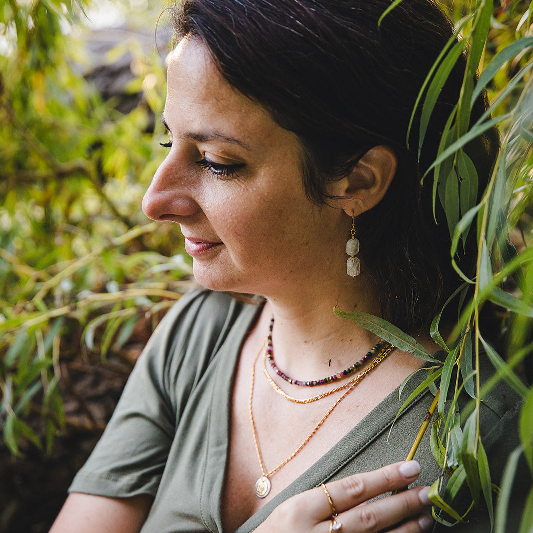 Femme portant les boucles d’oreilles Phoebe en pierre de lune arc-en-ciel et acier inoxydable doré, photo en extérieur dans un décor naturel.