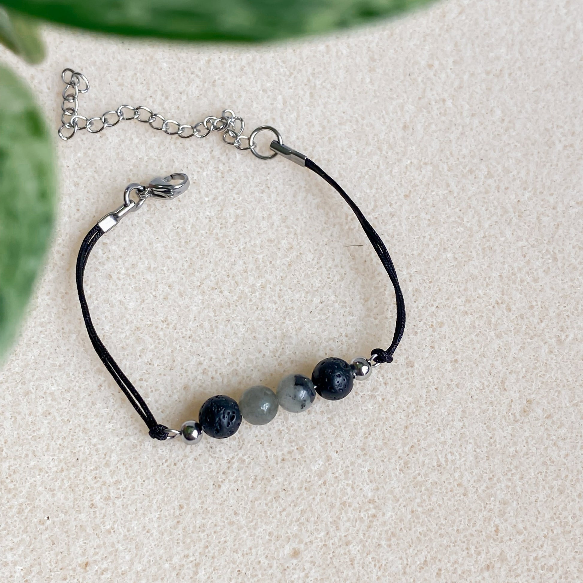 Bracelet ajustable en pierre volcanique et labradorite, cordon noir et fermoir argent, posé sur une surface texturée beige.
