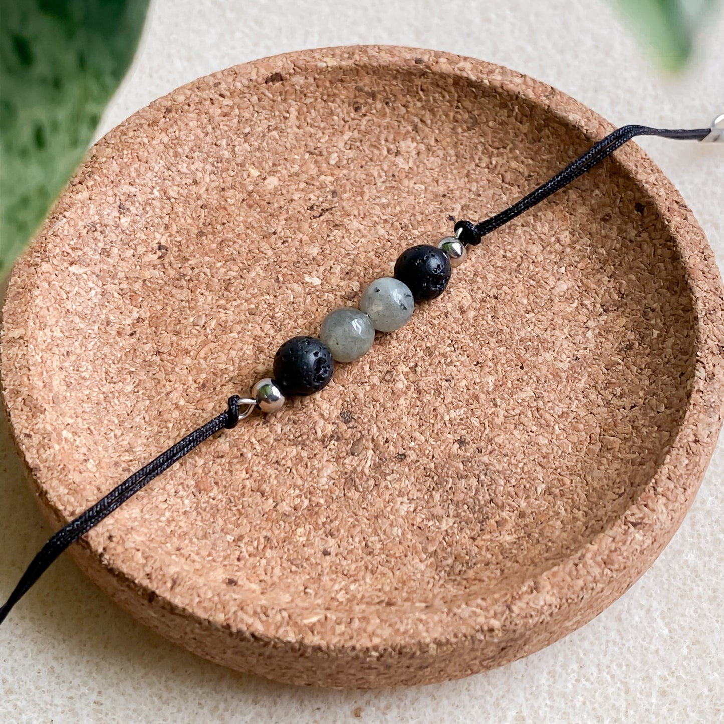 Bracelet cordon noir avec pierres naturelles de lave et labradorite, présentation plate sur un socle rond en liège.