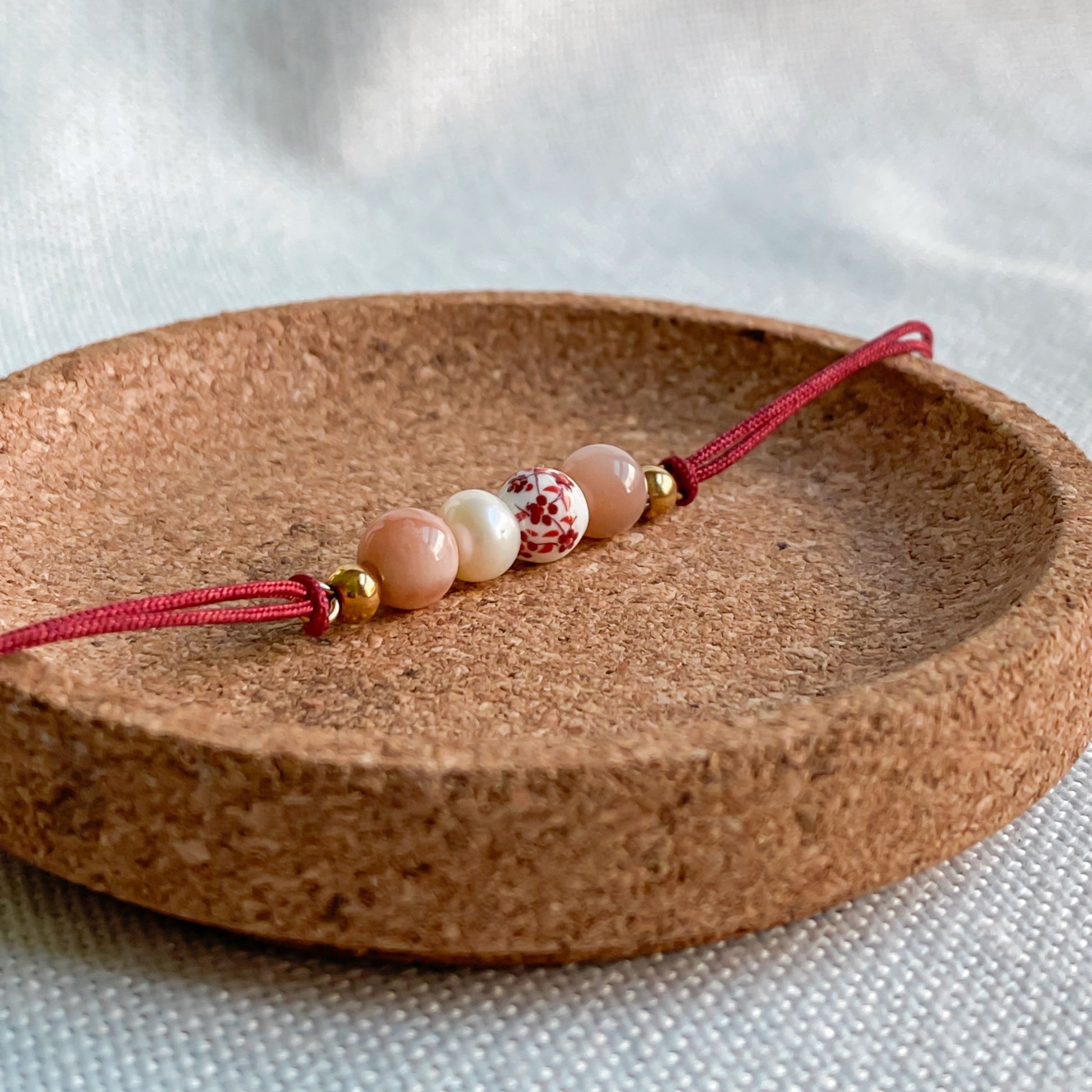 Bracelet cordon rouge en pierres naturelles Éveil présenté en gros plan sur support en liège, avec pierre de soleil, perle d’eau douce et céramique fleurie.