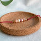Bracelet cordon rouge ajustable Éveil vu de dessus, bijou artisanal fait main à Montréal avec pierre de soleil, perle d’eau douce et céramique blanche fleurie.