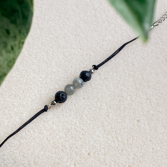 Bracelet diffuseur d’huiles essentielles en pierre de lave et labradorite, photographie minimaliste sur fond clair avec feuille verte.