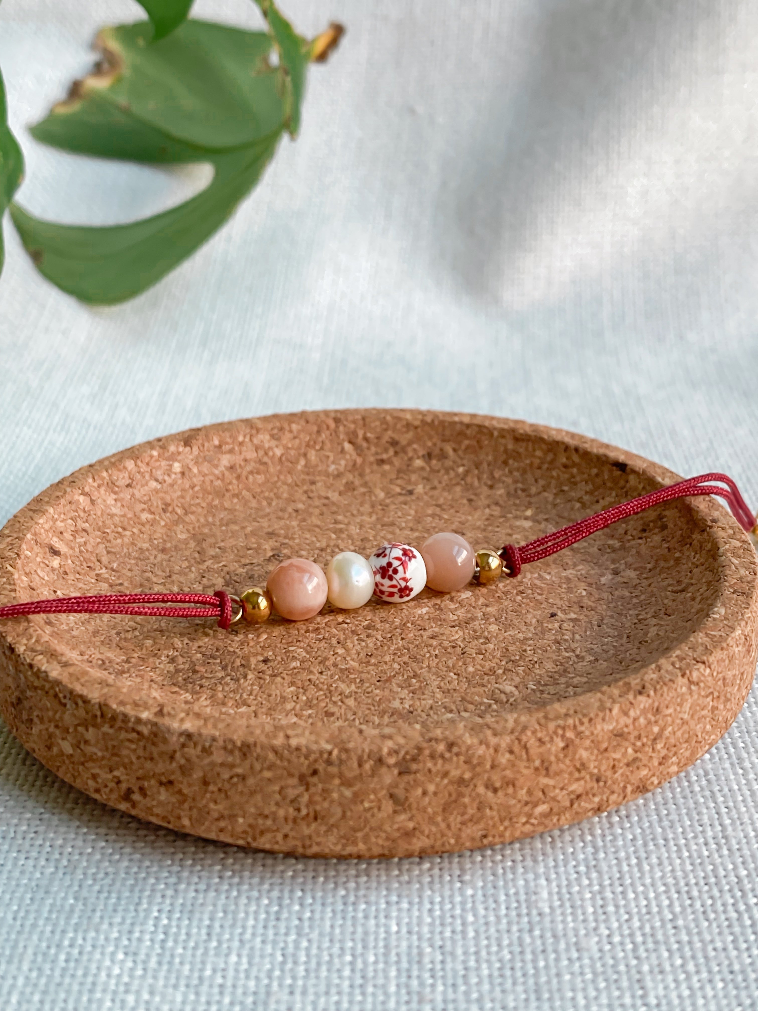 Bracelet cordon à personnaliser Doux Souvenir avec cordon rouge, bille de céramique blanche fleurie, perle d’eau douce et pierre de soleil, finition dorée.