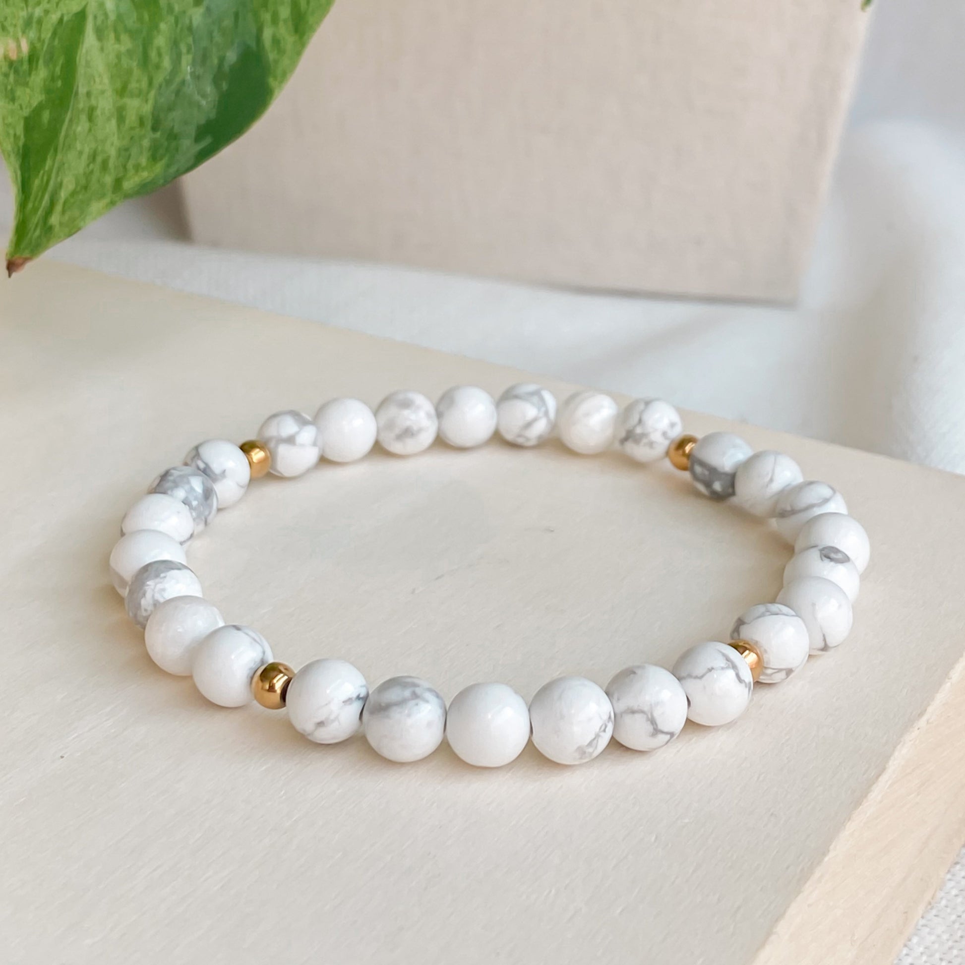 Bracelet élastique en howlite 6mm et acier inoxydable doré, légèrement vu de côté, posé sur une planche de bois clair avec une nappe en coton naturel et des plantes autour.