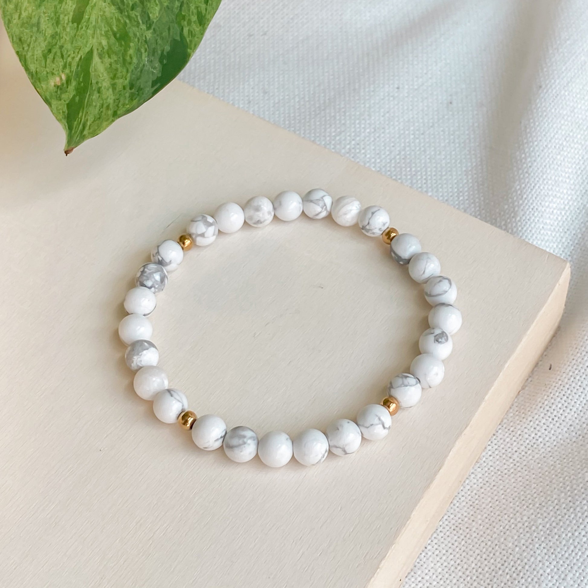 Bracelet en howlite 6mm et acier inoxydable doré, vu légèrement de côté, posé sur une planche de bois clair avec une nappe en coton naturel et des plantes autour.