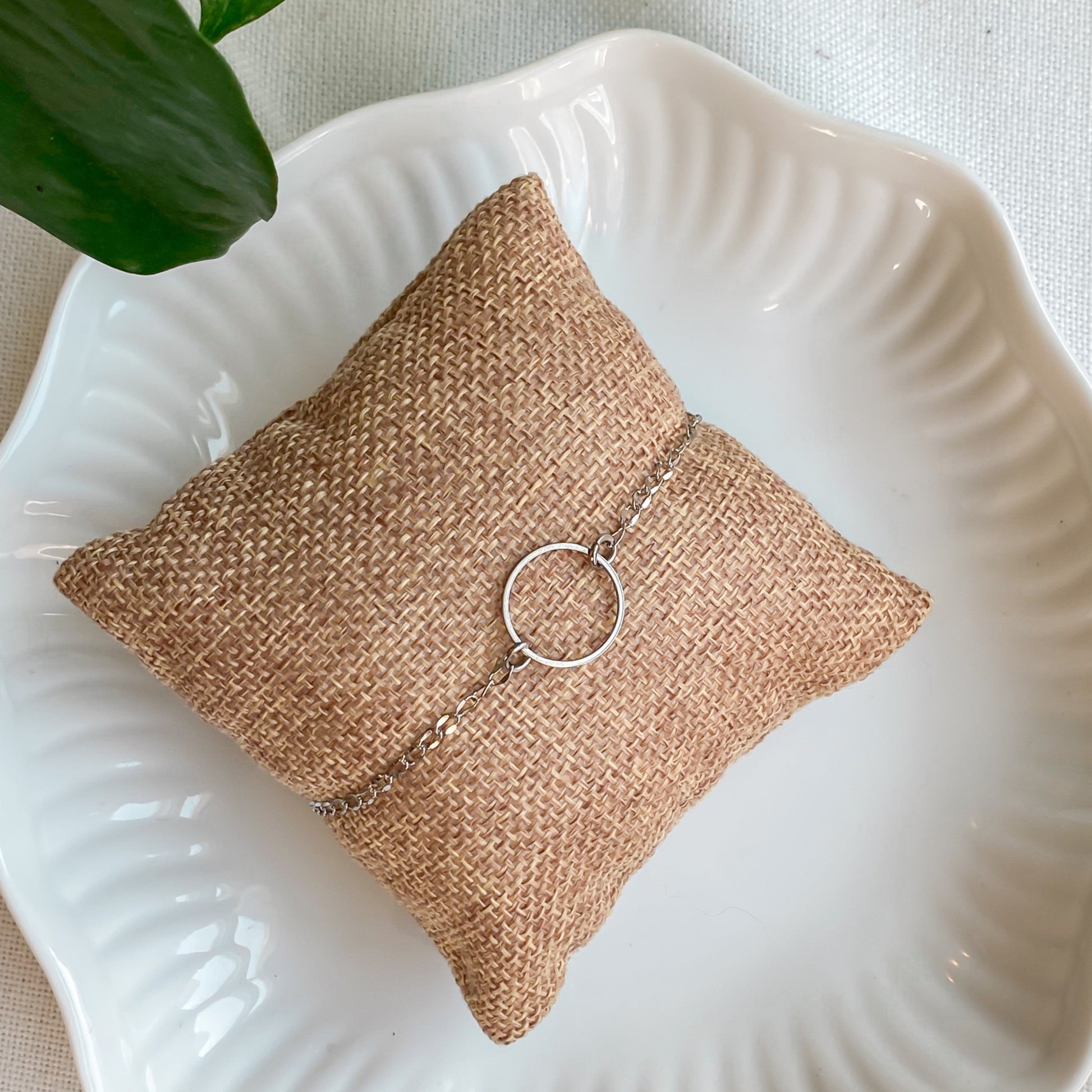 Bracelet minimaliste en chaîne fine en acier inoxydable avec un cercle, modèle Cernes, vue de haut sur un coussin en lin avec des feuilles de plantes.