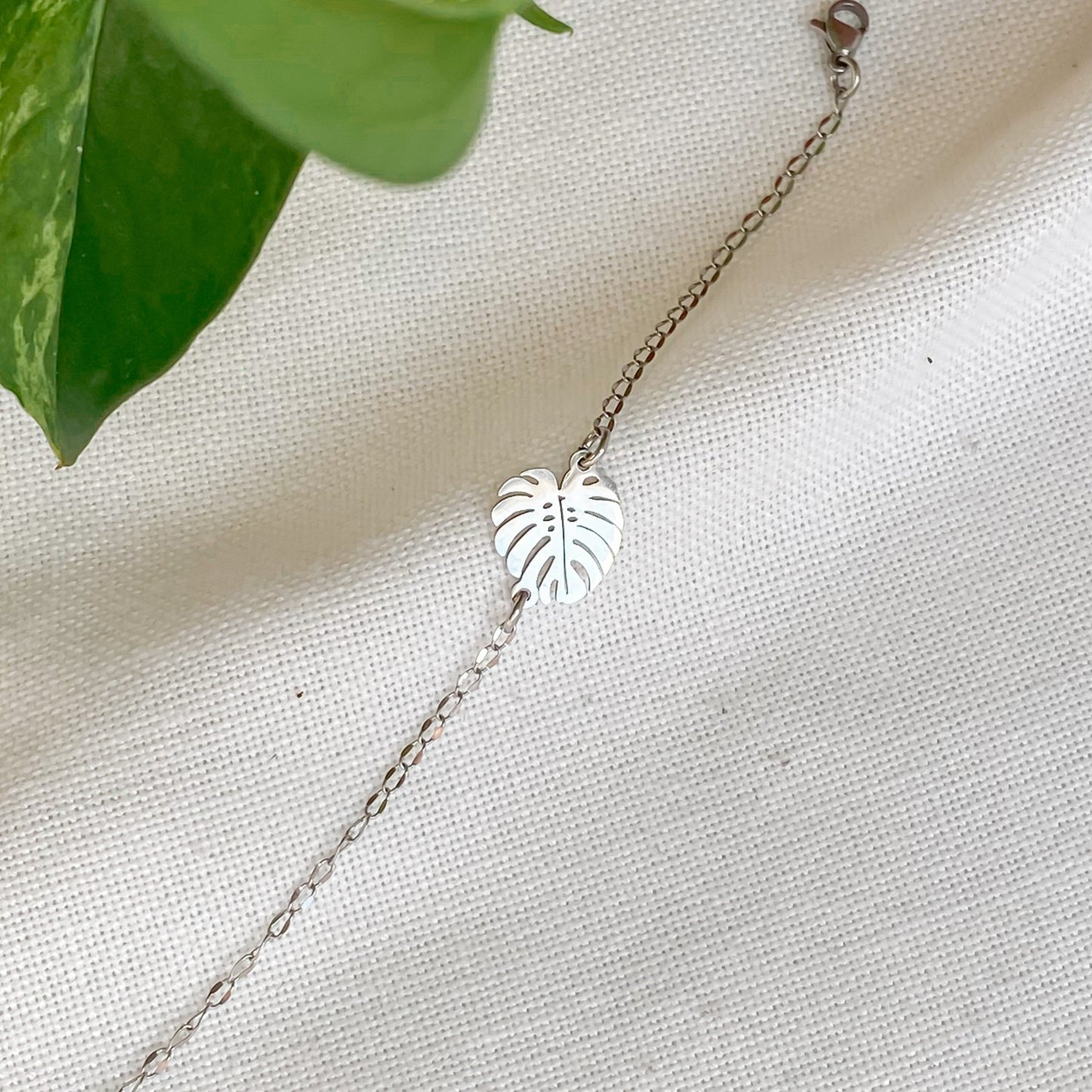 Bracelet minimaliste en chaîne fine avec pendentif feuille de monstera, posé sur un tissu blanc cassé avec des feuilles de plantes en arrière-plan.