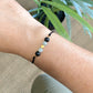 Bracelet NOCTIS pour aromathérapie en pierre de lave et labradorite, cordon noir fin et ajustable, porté au poignet avec plante verte en arrière-plan.