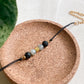 Bracelet artisanal NOCTIS diffuseur d’aromathérapie, cordon noir et pierres naturelles, présenté sur support rond en liège.