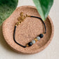 Bracelet NOCTIS diffuseur d’huiles essentielles en pierre de lave et labradorite, cordon noir avec fermoir doré, présenté sur support en liège.