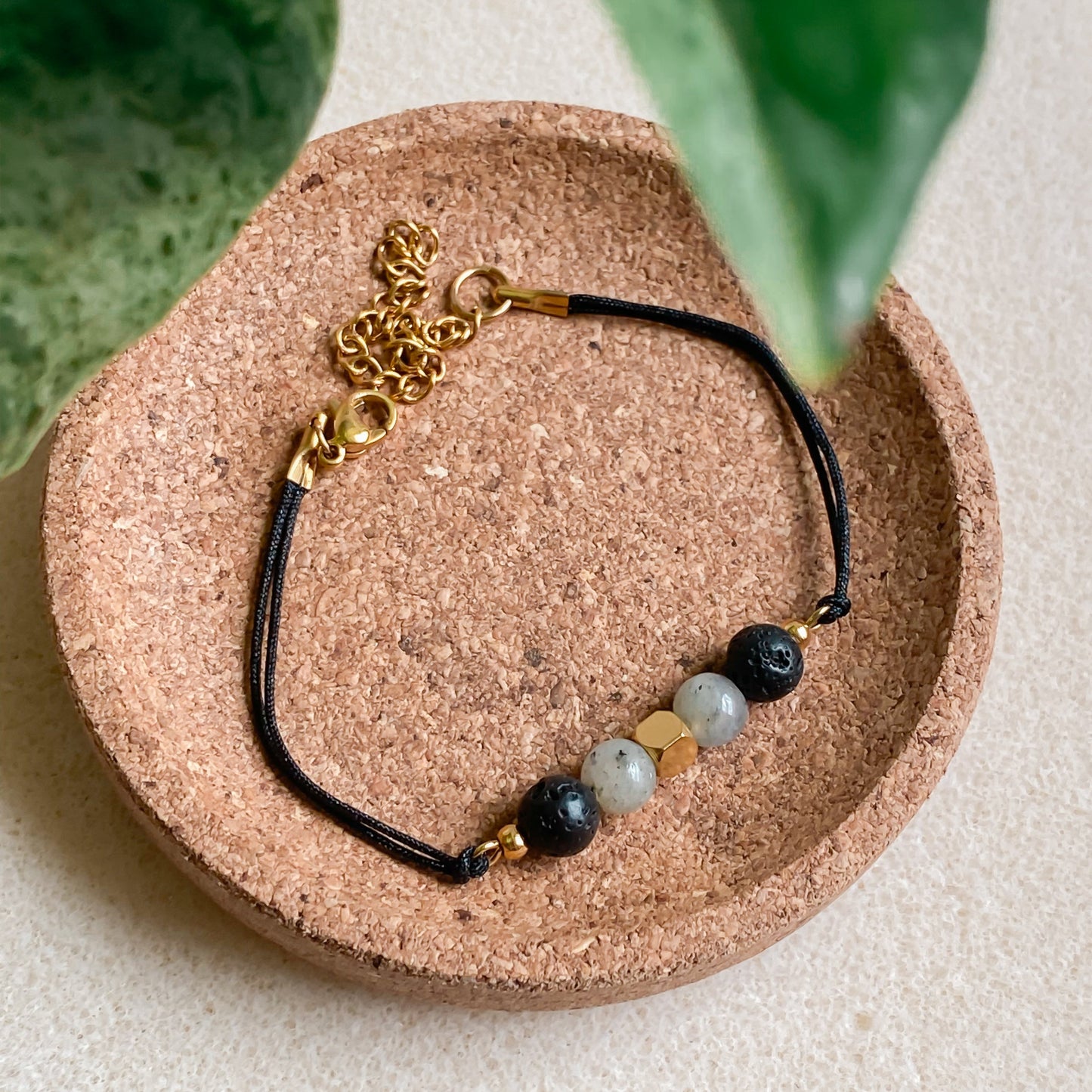 Bracelet NOCTIS diffuseur d’huiles essentielles en pierre de lave et labradorite, cordon noir avec fermoir doré, présenté sur support en liège.