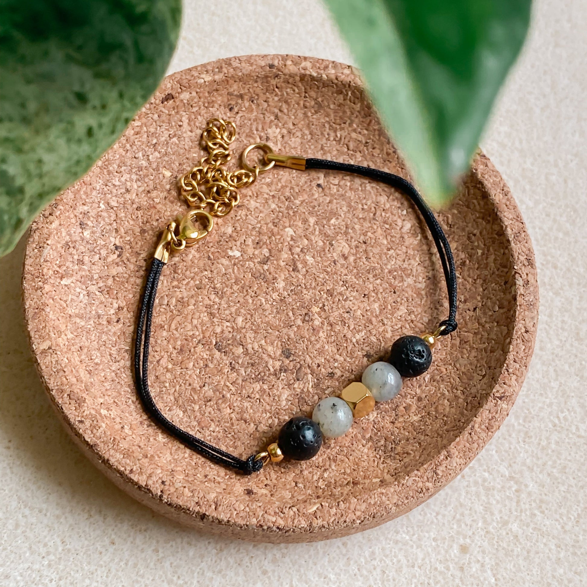 Bracelet NOCTIS diffuseur d’huiles essentielles en pierre de lave et labradorite, cordon noir avec fermoir doré, présenté sur support en liège.