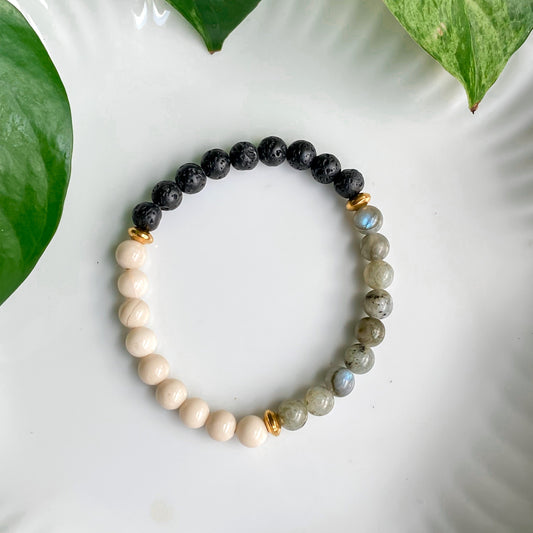 Bracelet élastique diffuseur d'huiles essentielles Sillans en lave noire, labradorite et pierre de rivière, avec acier inoxydable doré, présenté sur fond blanc entouré de plantes.